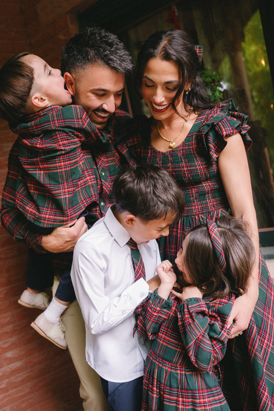 A family of five, all smiling, pose together. The mother and three children wear matching Ivy City Co’s Hattie Dress in Evergreen Plaid—perfect for the season. The father wears a plaid shirt and holds one child as they laugh. The mother’s stylish, maternity-friendly Hattie Dress ensures comfort while beautifully coordinating with her family’s festive look.