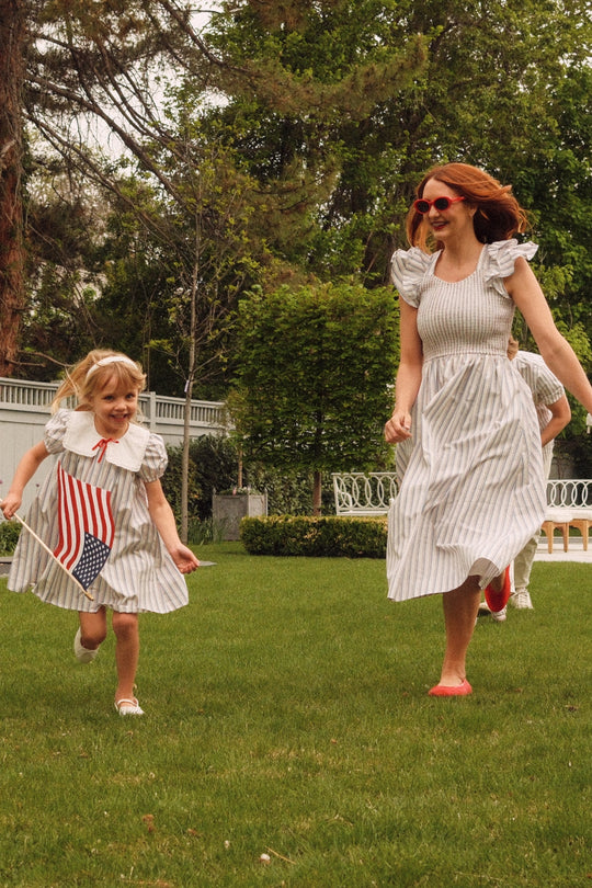 A woman and a young girl, both wearing the Hattie Dress in Heritage Stripe by Ivy City Co, run joyfully across green grass. The girl waves an American flag as trees and bushes form a lively backdrop.