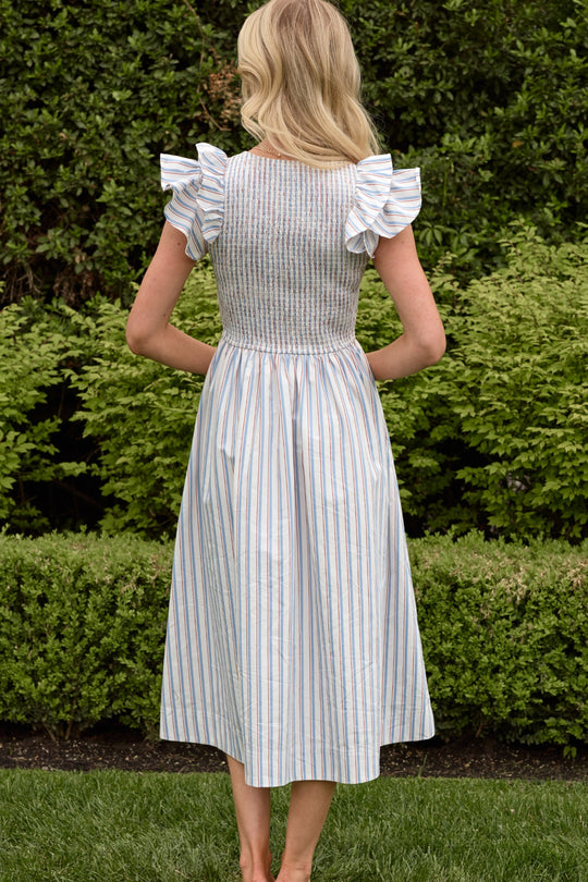 A woman with blonde hair stands outdoors facing away, wearing the Ivy City Co Hattie Dress in Heritage Stripe, featuring vertical pastel stripes and ruffled sleeves. Green bushes and grass are seen in the background.