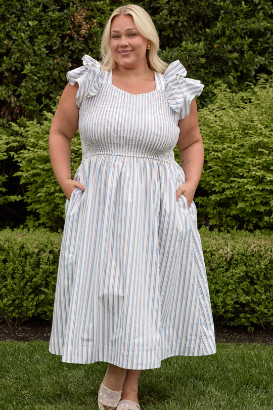 A woman with blonde hair stands on grass, smiling with her hands in the pockets of the Ivy City Co Hattie Dress in Heritage Stripe, featuring blue and pink vertical stripes and ruffled sleeves. Green bushes form the backdrop.