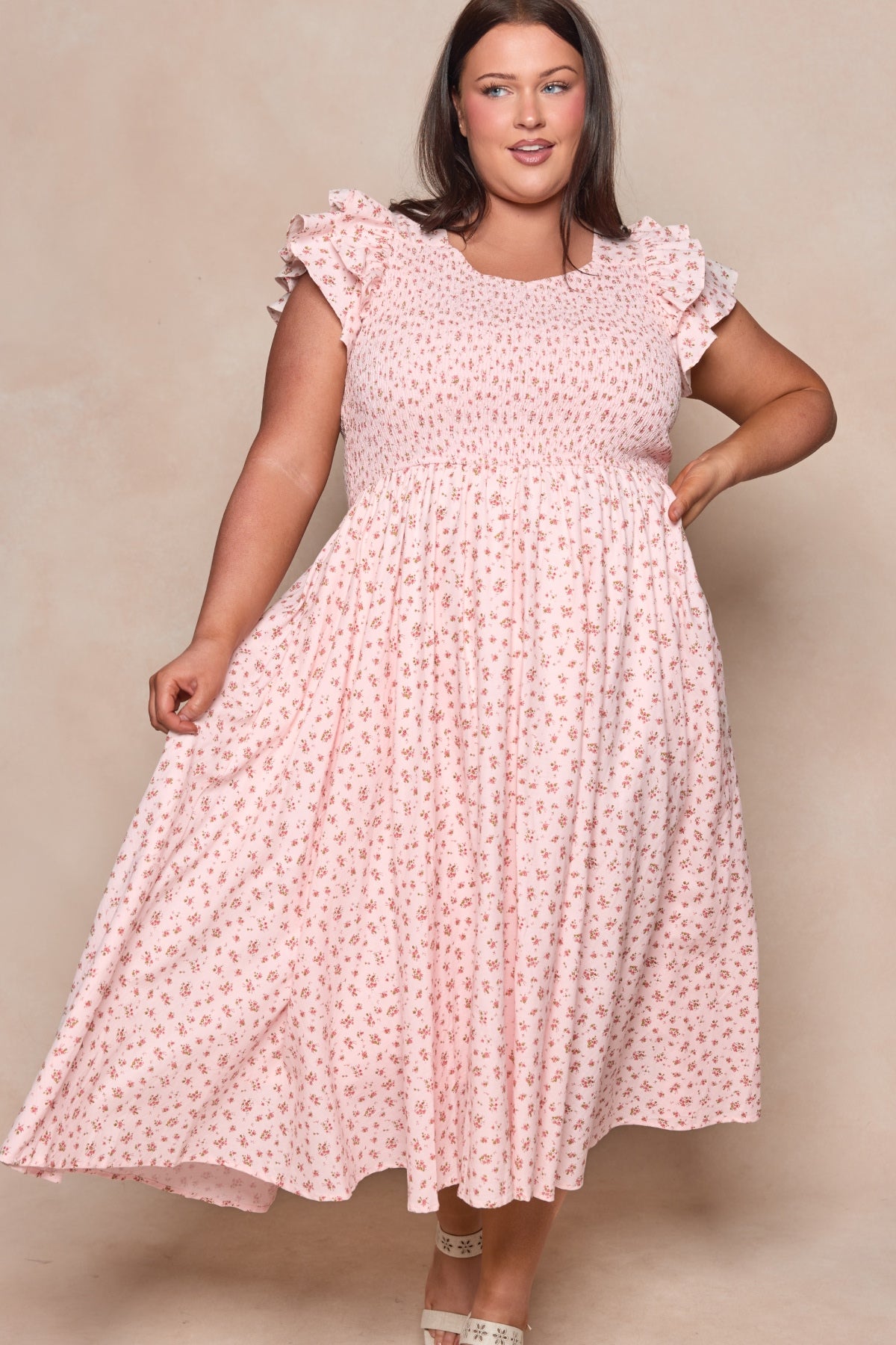 A woman in the Hattie Dress in Madeline Pink by Ivy City Co—a light pink, short-sleeved cotton midi dress with a small floral print—stands against a neutral backdrop, smiling and holding out her dress. A chic choice for a birthday girl.