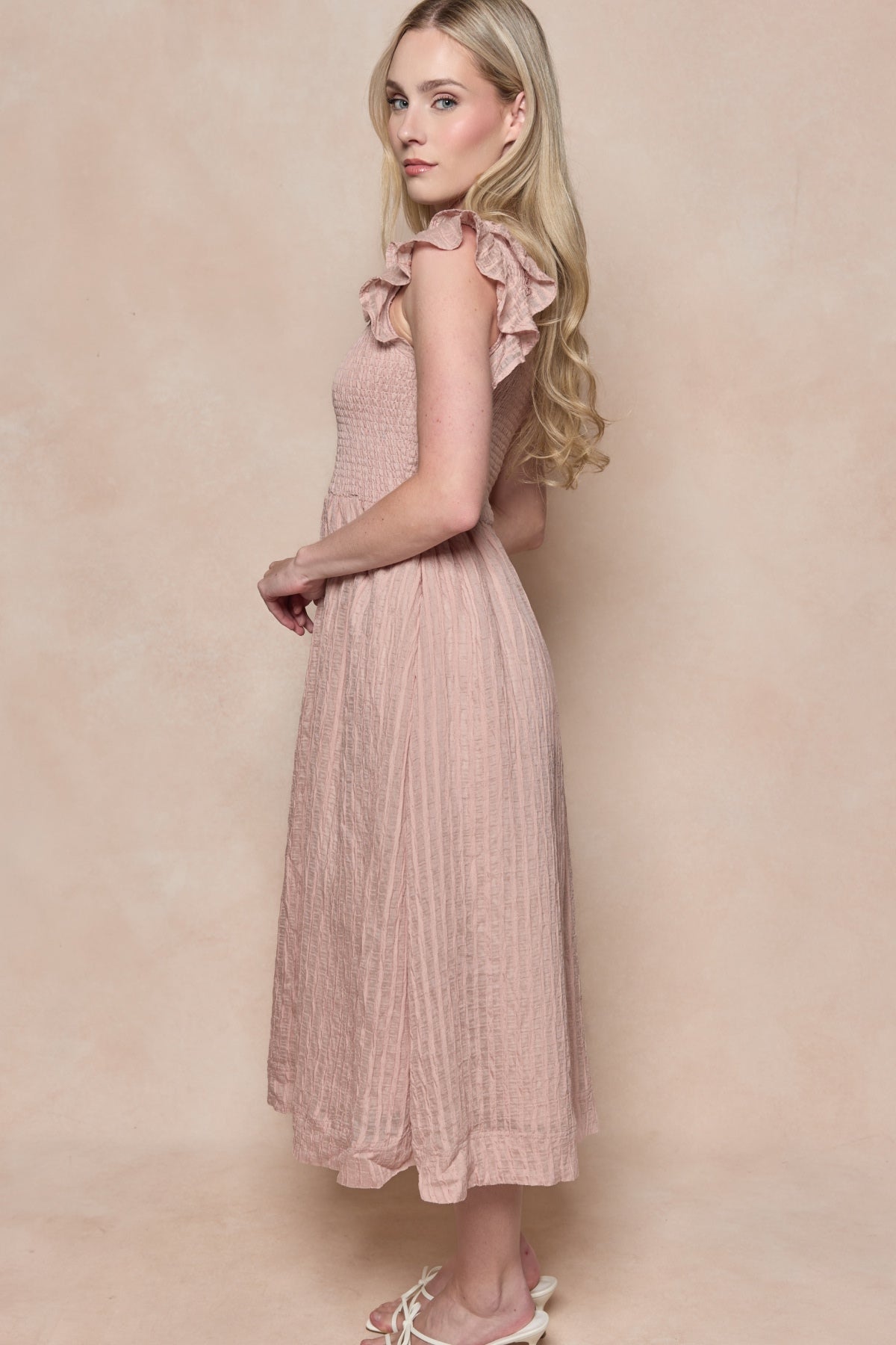A woman with long blonde hair stands sideways in the Hattie Dress in Pink Stripes by Ivy City Co, featuring ruffle details and a smocked bodice, paired with white sandals against a soft beige background.