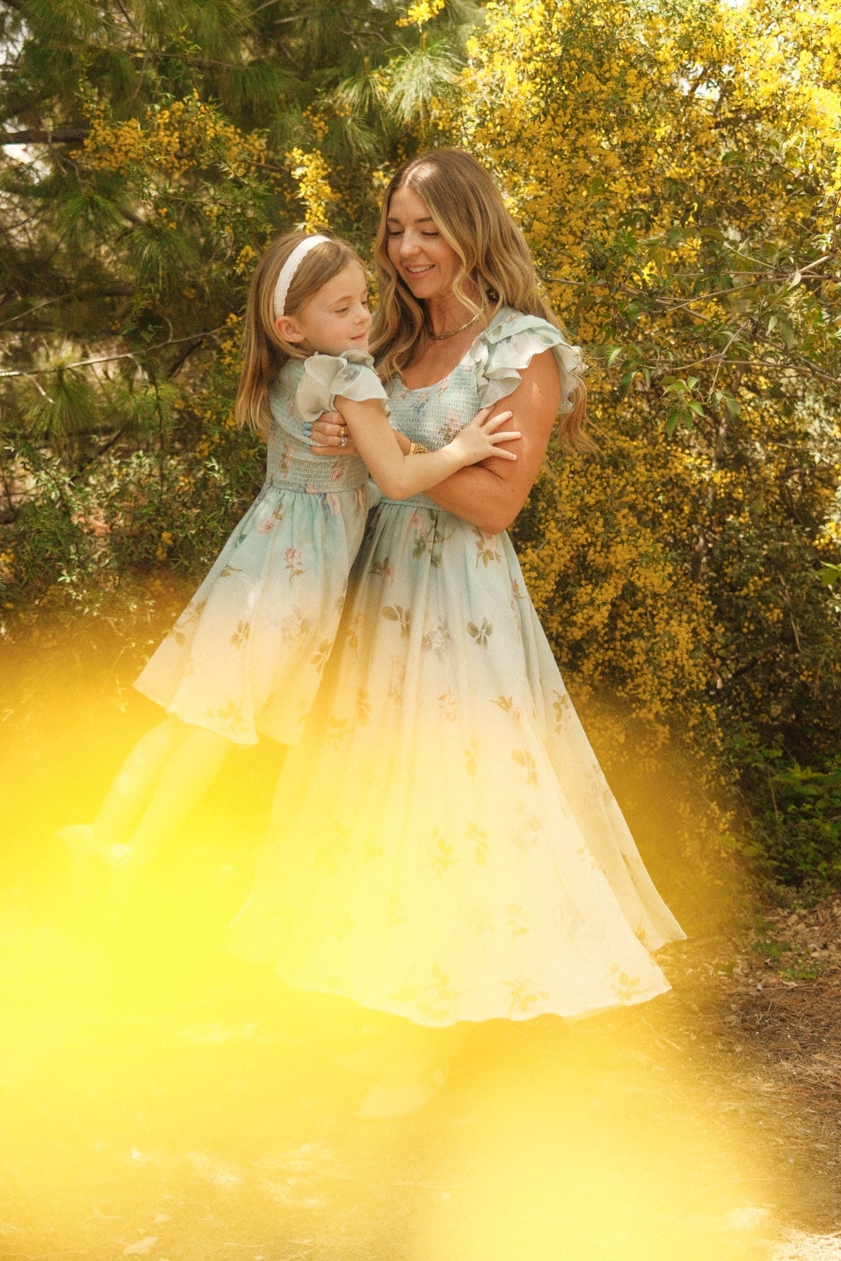 A woman and a young girl wear matching Ivy City Co Hattie Dresses in Roselyn Blue, smiling together amid yellow flowers and greenery as sunlight bathes the scene in a warm, golden glow.