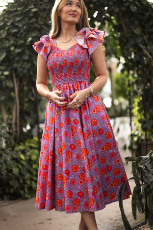 A woman stands outdoors in a garden, wearing the Ivy City Co Hattie Dress in Thistle Flower Poppy Woodblock—a purple floral, maternity-friendly dress with ruffled sleeves—her hands clasped and smiling gently as she looks upward.
