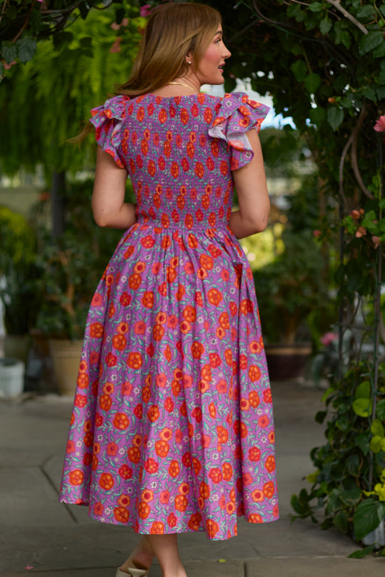A woman stands outdoors among lush greenery wearing Ivy City Co’s Hattie Dress in Thistle Flower Poppy Woodblock—a vibrant, maternity-friendly purple floral dress with ruffled cap sleeves, perfect for sunny days.