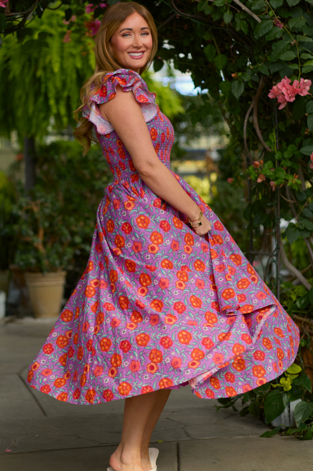 A woman with long hair smiles at the camera while twirling outdoors in lush greenery and pink flowers, wearing the Ivy City Co Hattie Dress in Thistle Flower Poppy Woodblock, a maternity-friendly floral dress.