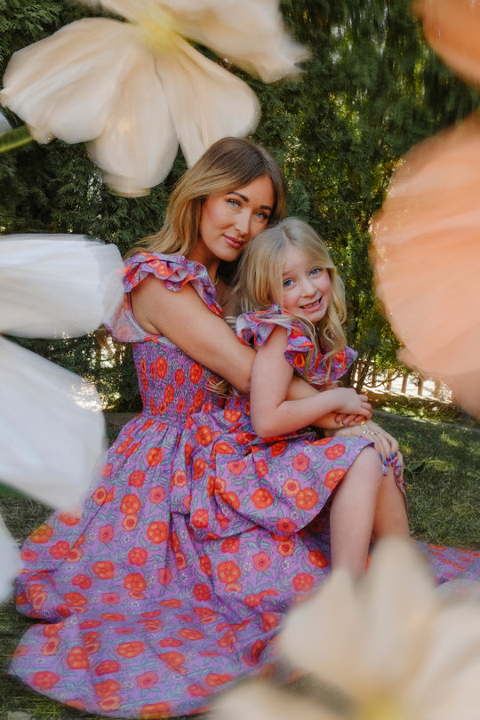 A woman and young girl sit on the grass, hugging in matching Ivy City Co Hattie Dress in Thistle Flower Poppy Woodblock—perfect as a nursing-friendly or maternity-friendly dress, surrounded by softly blurred flowers and greenery.