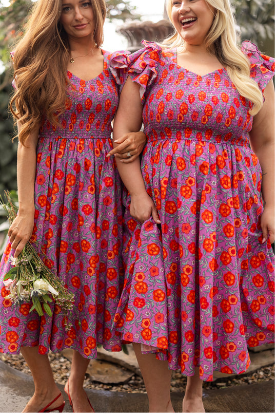 Two women stand outdoors, smiling in matching Ivy City Co Hattie Dresses in Thistle Flower Poppy Woodblock—nursing-friendly floral dresses with ruffled sleeves, perfect for joyful moments together. One woman holds a bouquet of flowers.