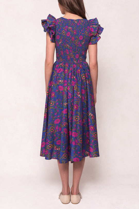 A woman stands facing away, wearing the Ivy City Co Hattie Dress in Blue Multicolor Floral, featuring a smocked bodice, ruffled short sleeves, and a vibrant floral pattern that’s both stylish and maternity friendly.