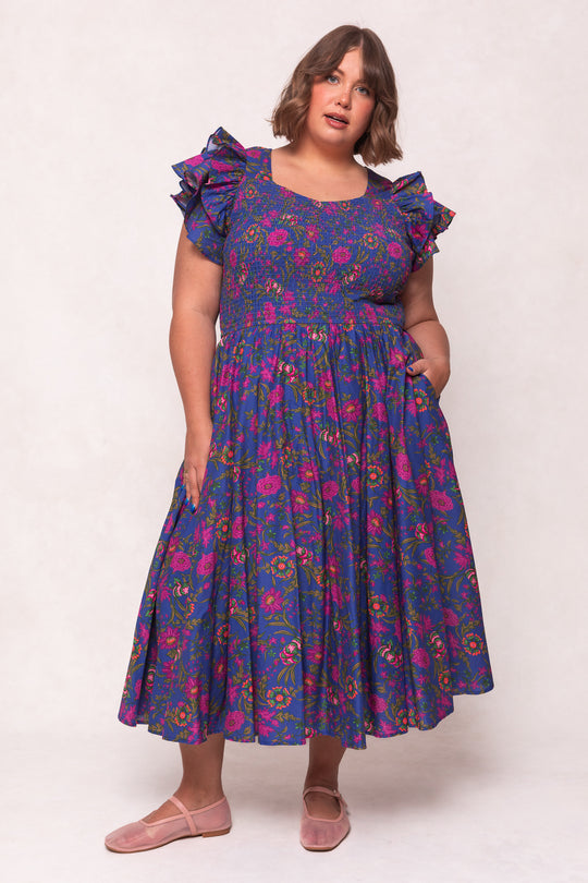 A woman with shoulder-length brown hair and pink flats stands against a light background, wearing the Ivy City Co Hattie Dress in Blue Multicolor Floral. The cotton midi dress features short sleeves, ruffles, and pink flowers.