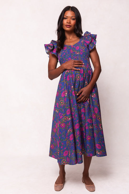 A woman wears the Ivy City Co Hattie Dress in Blue Multicolor Floral, featuring ruffled sleeves and a flowing cotton midi skirt, styled with long dark hair and light flat shoes against a light background for a chic look.