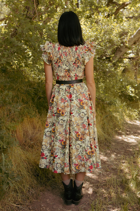 A woman with straight black hair stands on a forest path, wearing the Ivy City Co Hattie Dress in Rose Vine—featuring a smocked bodice, ruffled sleeves, and a maternity-friendly fit—styled with a black belt and boots amid lush greenery.