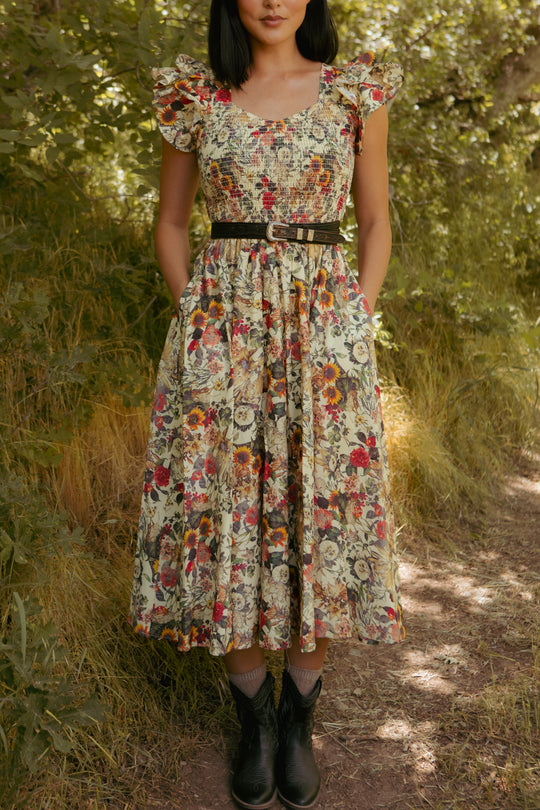 A woman stands outdoors on a dirt path wearing the Ivy City Co Hattie Dress in Rose Vine, with a smocked bodice, ruffled sleeves, black belt, and dark ankle boots. Her hands are in her pockets as sunlight streams through green foliage behind her.