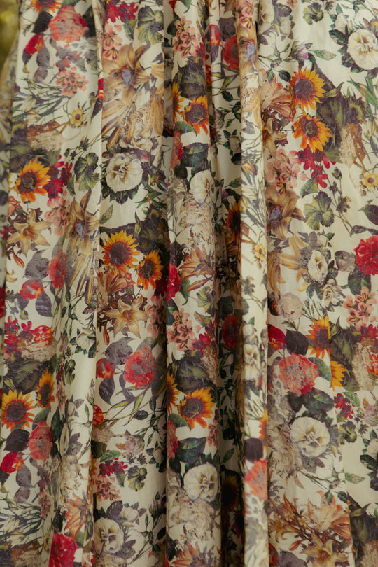 A close-up of Ivy City Co’s Hattie Dress in Rose Vine shows a cream fabric densely covered with red, yellow, orange, white, and purple florals and green leaves—ideal for its smocked, maternity-friendly bodice.