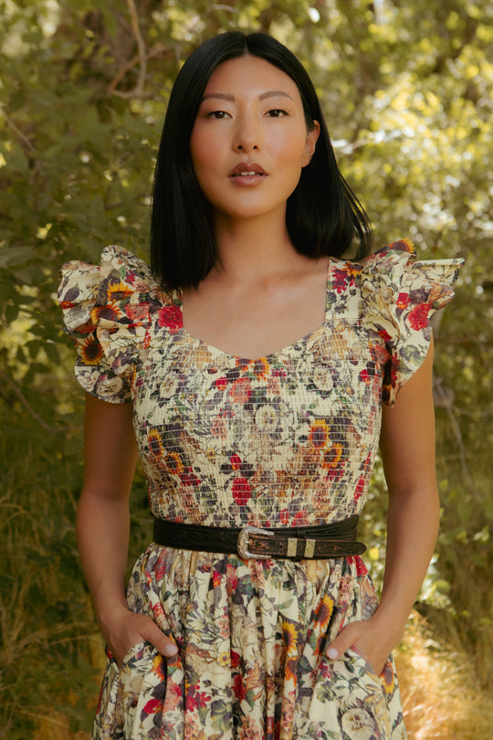 A woman with straight black hair stands outdoors in the Ivy City Co Hattie Dress in Rose Vine, featuring ruffled sleeves, a smocked bodice, a black belt, and pockets. Sunlight filters through greenery—maternity friendly and stylish.