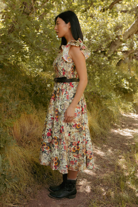 A woman stands sideways on a wooded path in the Hattie Dress in Rose Vine by Ivy City Co, a maternity-friendly floral piece with a smocked bodice, styled with a black belt and boots as sunlight filters through the trees.