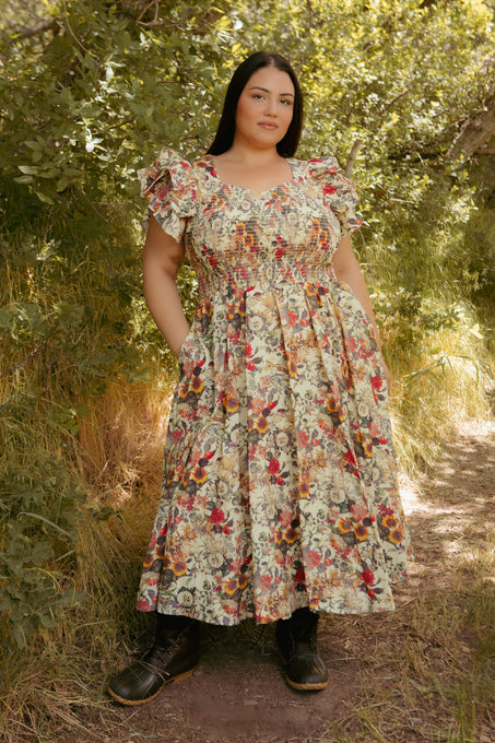 A woman stands outdoors on a sunlit path in the Ivy City Co Hattie Dress in Rose Vine, featuring a smocked bodice and pockets. Styled with black boots, her maternity-friendly look is effortlessly chic.