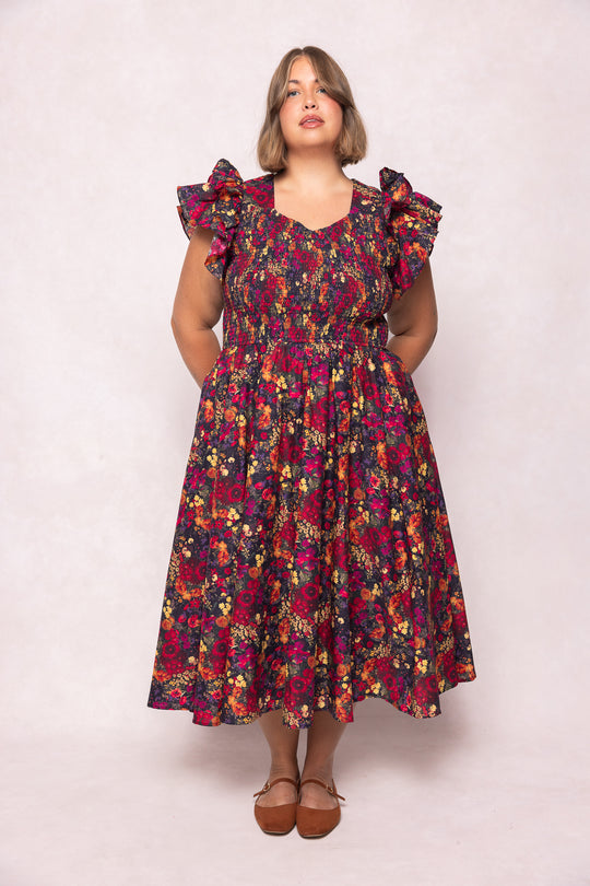 A woman with short light brown hair wears the Ivy City Co Hattie Dress in Night Garden, a dark floral midi with ruffled sleeves and a smocked bodice. She pairs it with brown flats and rests her hands in the dress pockets for a relaxed look.