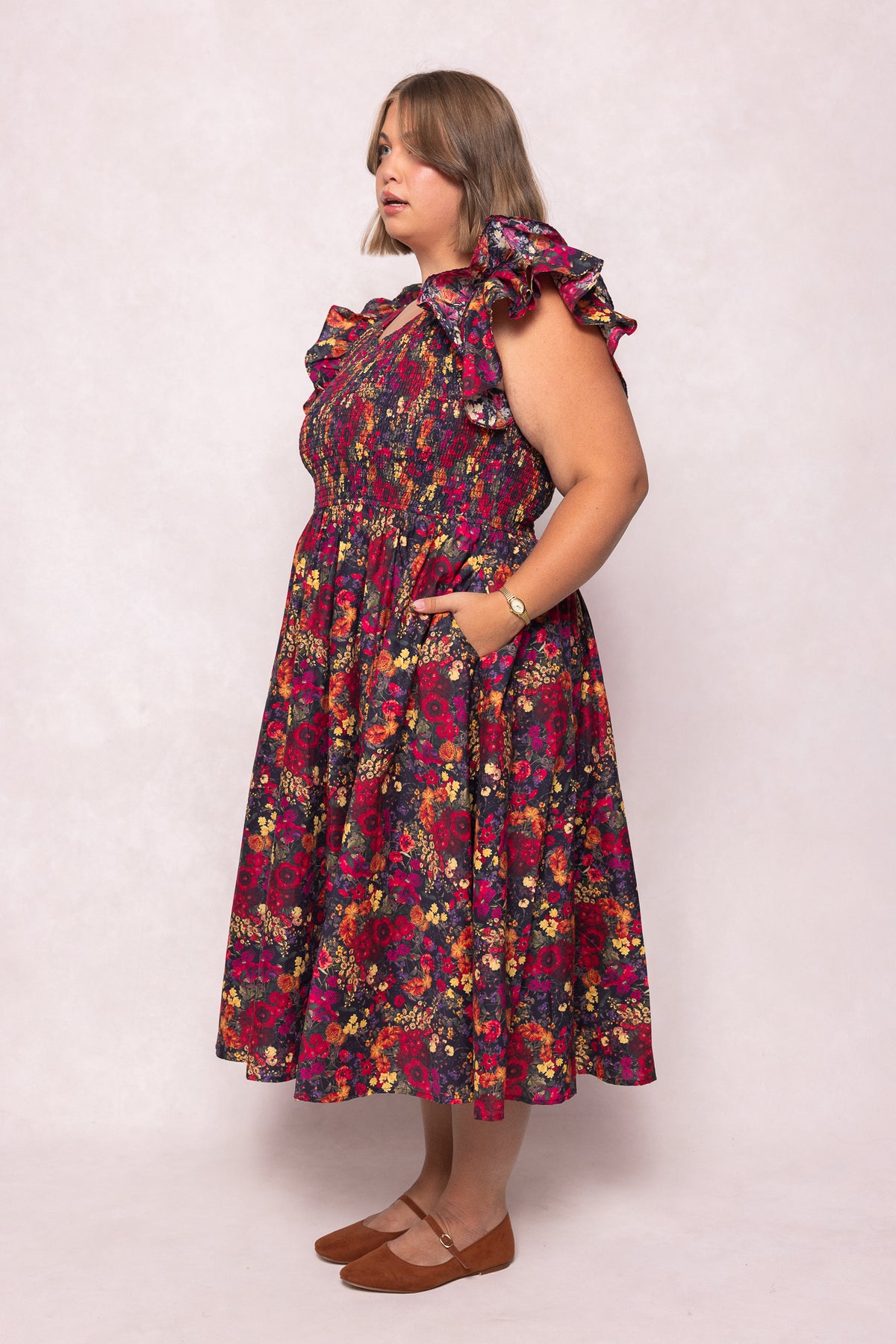 A woman stands in profile against a light background wearing the Ivy City Co Hattie Dress in Night Garden, a chic, maternity-friendly dark floral midi with smocked bodice and pockets. She has shoulder-length hair, brown flats, and a watch.