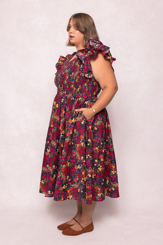 A woman stands in profile against a light background wearing the Ivy City Co Hattie Dress in Night Garden, a chic, maternity-friendly dark floral midi with smocked bodice and pockets. She has shoulder-length hair, brown flats, and a watch.