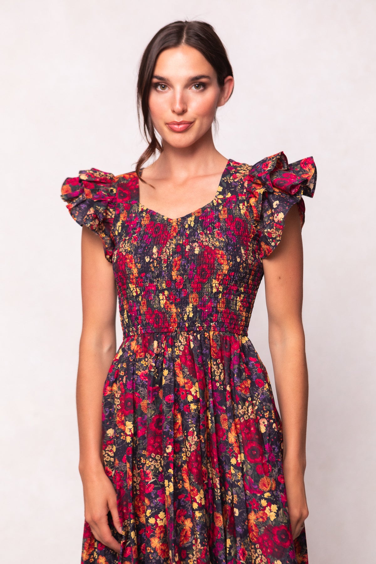 A woman wears the Ivy City Co Hattie Dress in Night Garden, featuring a dark floral print, ruffled cap sleeves, and a smocked bodice, standing against a light background and smiling slightly at the camera.