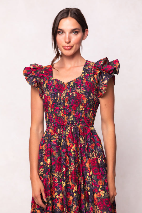 A woman wears the Ivy City Co Hattie Dress in Night Garden, featuring a dark floral print, ruffled cap sleeves, and a smocked bodice, standing against a light background and smiling slightly at the camera.