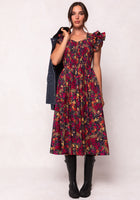 A woman faces forward in the Ivy City Co Hattie Dress in Night Garden—a dark floral midi with puffed sleeves and a smocked bodice—styled with black knee-high boots and a dark jacket over her shoulder against a light background.