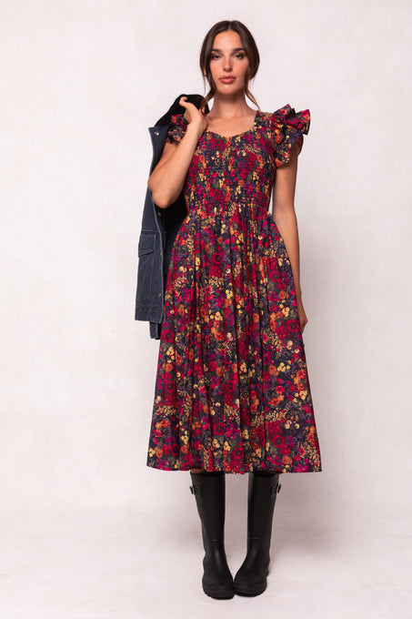A woman faces forward in the Ivy City Co Hattie Dress in Night Garden—a dark floral midi with puffed sleeves and a smocked bodice—styled with black knee-high boots and a dark jacket over her shoulder against a light background.