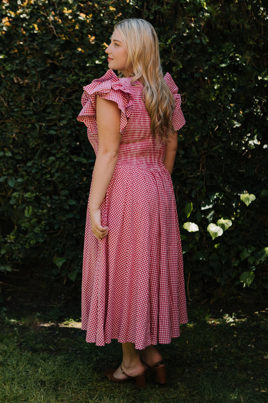 A woman with long blonde hair stands outdoors in the Ivy City Co Hattie Dress in Red Gingham, featuring ruffled sleeves and a smocked bodice. She pairs it with brown heeled sandals, set against lush green foliage.