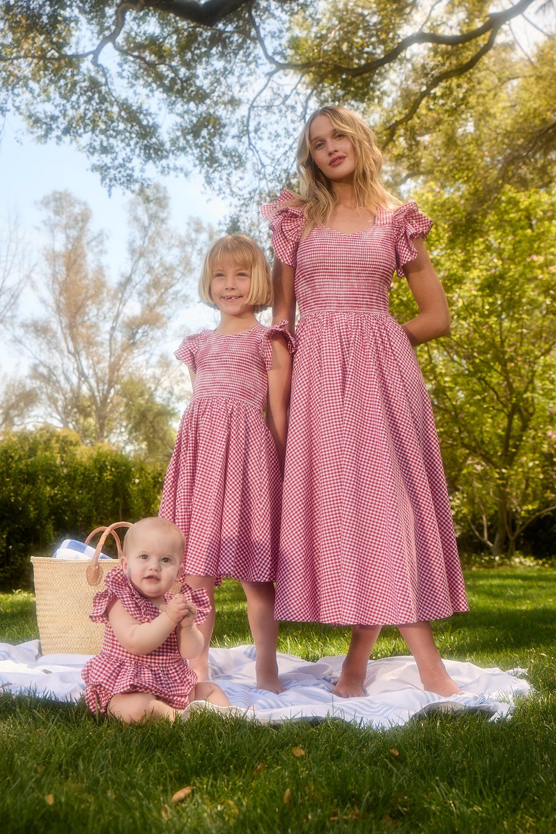 A woman, a young girl, and a baby in Ivy City Co’s Baby Hattie Romper in Red Gingham and matching dresses relax on a blanket outdoors, with a picnic basket nearby among trees—a charming scene for toddler clothing.