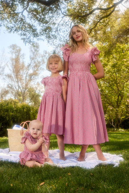 A woman, a young girl, and a baby in Ivy City Co’s Baby Hattie Romper in Red Gingham and matching dresses relax on a blanket outdoors, with a picnic basket nearby among trees—a charming scene for toddler clothing.