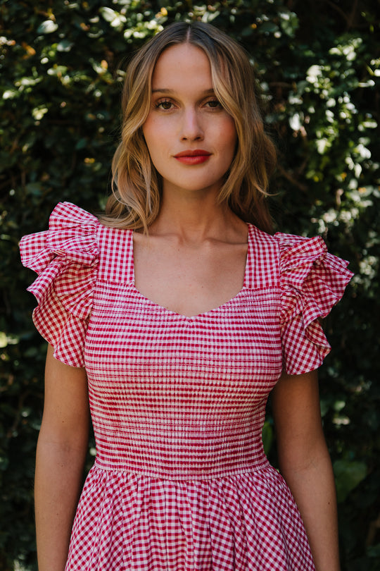 A woman stands outdoors before leafy greenery, wearing the Ivy City Co Hattie Dress in Red Gingham—featuring a smocked bodice and ruffled sleeves—showcasing this stylish, maternity-friendly look.