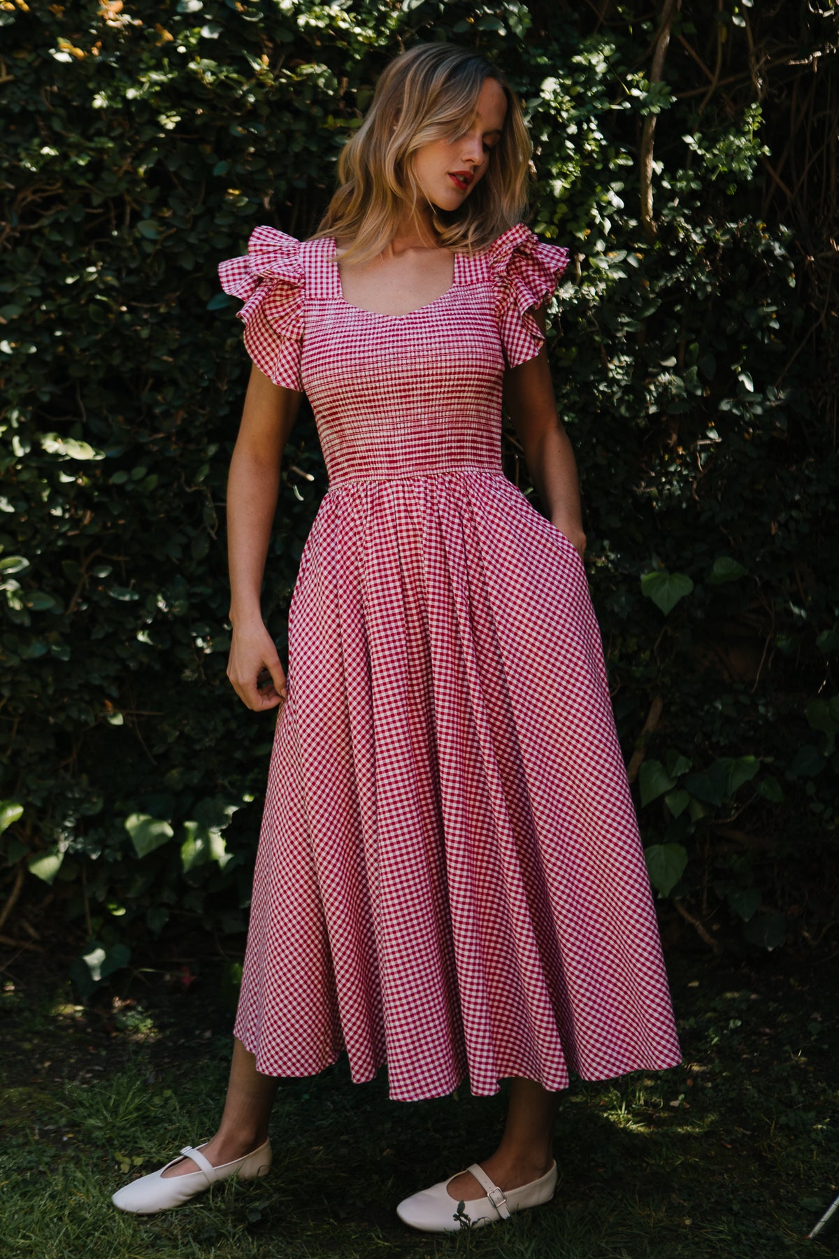A woman stands outdoors before leafy bushes, wearing Ivy City Cos Hattie Dress in Red Gingham with ruffled sleeves, a smocked bodice, and full skirt. Looking down to her left, she completes the look with white shoes.