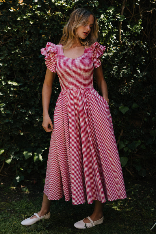 A woman stands outdoors before leafy bushes, wearing Ivy City Cos Hattie Dress in Red Gingham with ruffled sleeves, a smocked bodice, and full skirt. Looking down to her left, she completes the look with white shoes.