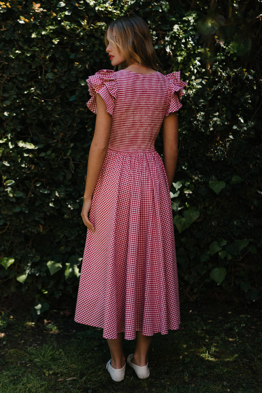 A woman stands outdoors among green bushes and grass, wearing the Ivy City Co Hattie Dress in Red Gingham. The dress has a smocked bodice, ruffled short sleeves, a partially open back, and she pairs it with light-colored shoes.