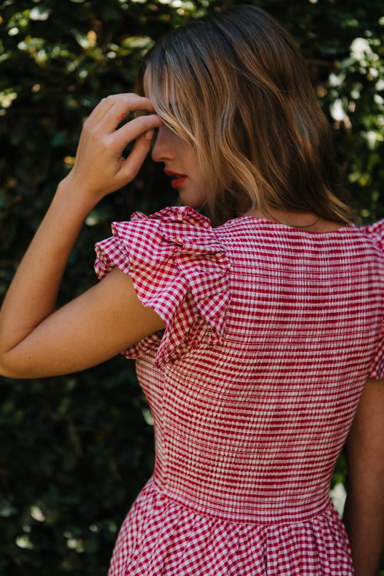 A woman stands outdoors, facing away and touching her hair, wearing the Ivy City Co Hattie Dress in Red Gingham with ruffled sleeves and a smocked bodice; greenery is softly blurred in the background.