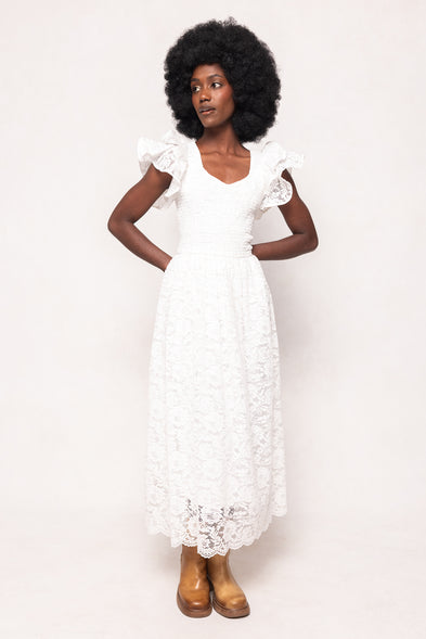 A woman with a large afro wears the Hattie Dress in White Lace by Ivy City Co—featuring ruffled shoulders—paired with brown ankle boots, standing against a plain background. This romantic midi dress is ideal for special occasions or bridal showers.