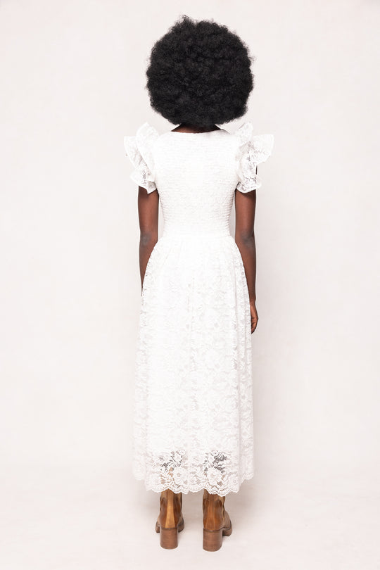 A person with a natural afro stands facing away, wearing the Ivy City Co Hattie Dress in White Lace with ruffled short sleeves and brown ankle boots, set against a plain light background.
