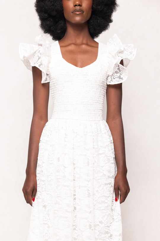 A woman with natural afro hair wears the Ivy City Co Hattie Dress in White Lace, featuring romantic ruffled shoulders and a fitted ruched bodice. Her hands are by her sides with red nails. The plain background highlights this perfect bridal shower dress.
