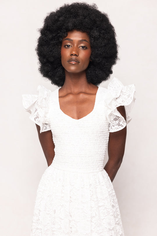 A woman with a natural afro wears the Ivy City Co Hattie Dress in White Lace, featuring romantic ruffled lace sleeves. She stands against a plain light background, confidently gazing at the camera with her hands behind her back.