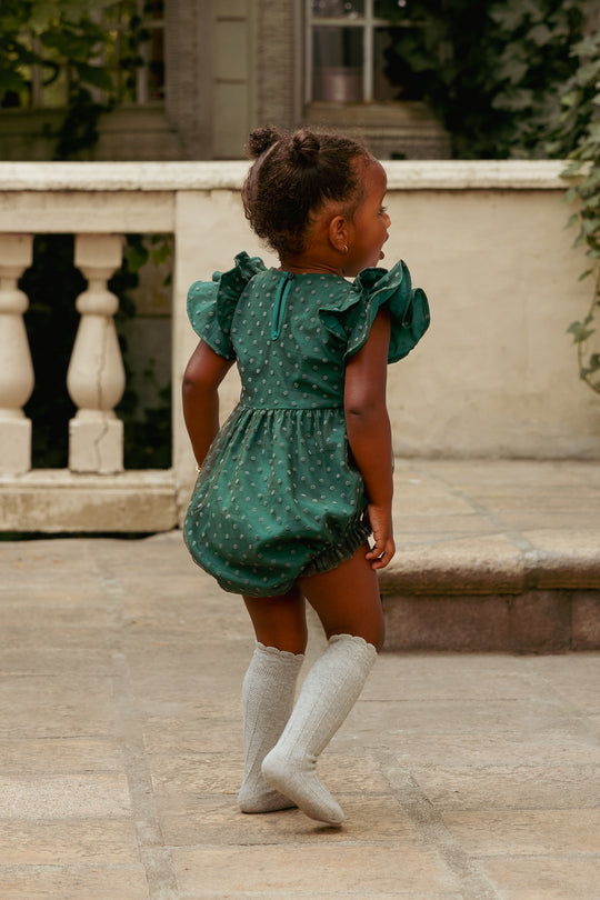 Child wearing a green polka dot romper with ruffled sleeves outdoors.