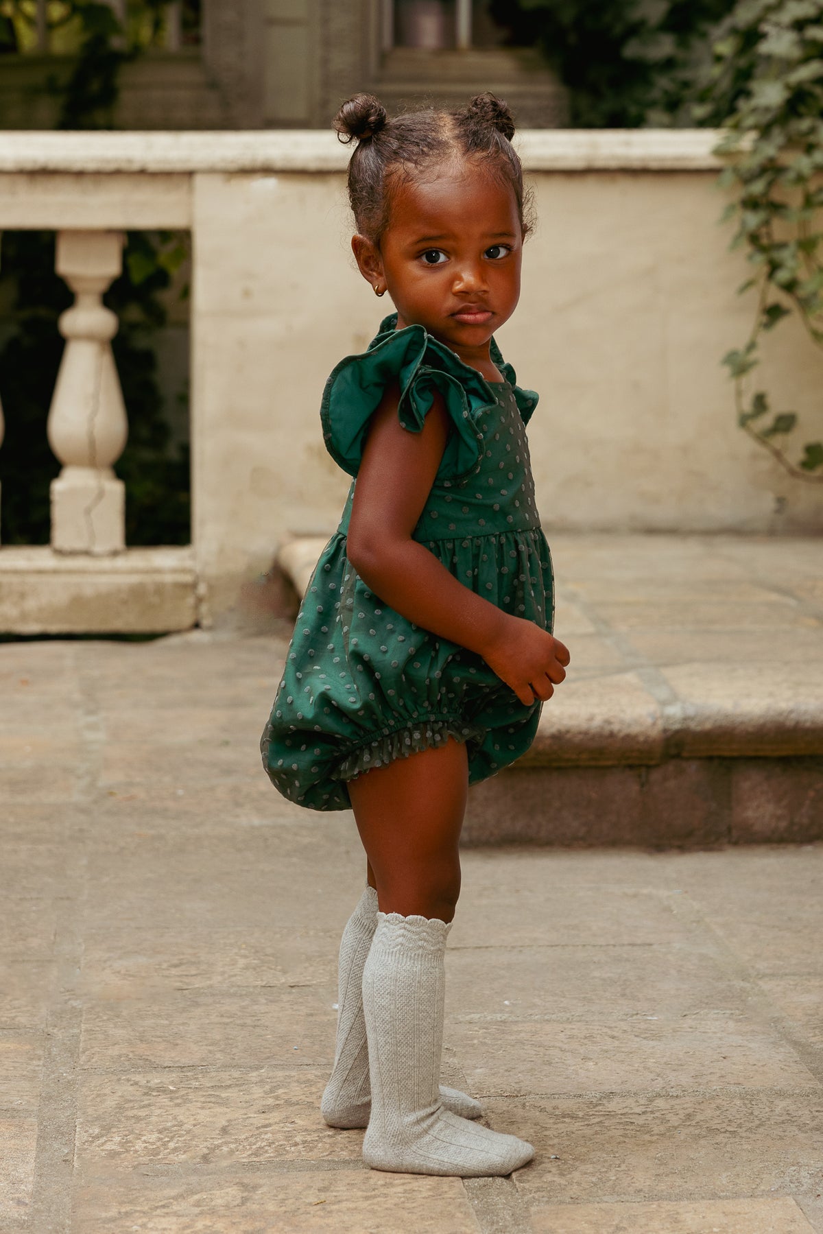 Young child wearing a green dress and white boots standing on a stone patio.