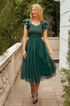 Woman in a green dress standing outdoors with greenery and a stone wall in the background