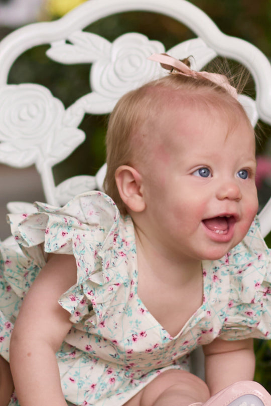 A smiling baby with light skin and blue eyes sits on a white floral chair, wearing Ivy City Co’s Baby Hattie Romper in Pansy Trellis as her Easter outfit, featuring ruffled sleeves and a small pink bow in her hair.