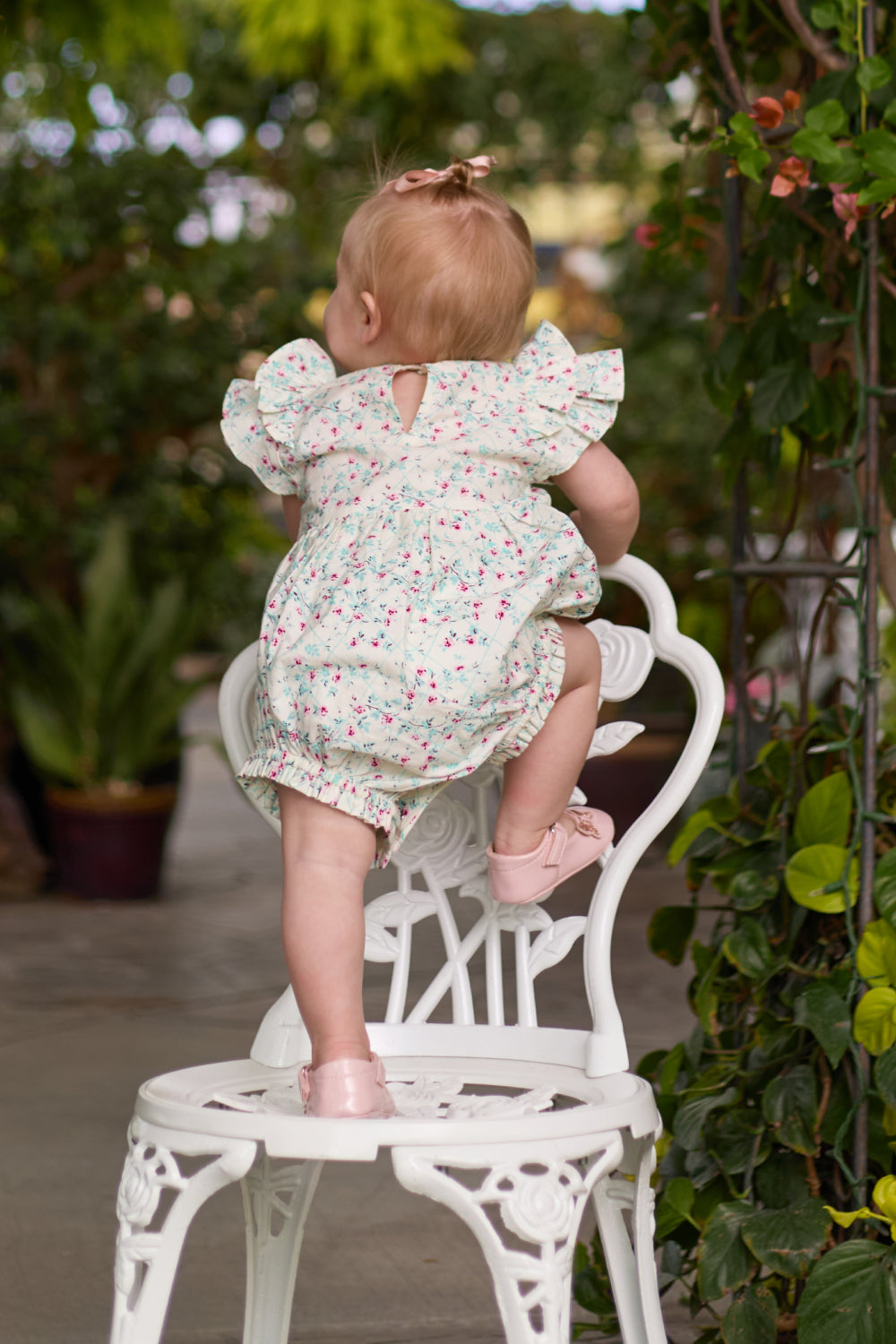 A toddler in the Ivy City Co Baby Hattie Romper in Pansy Trellis and pink shoes climbs onto a white ornate chair outdoors, green plants all around. Her floral romper stands out, with her back to the camera in this charming scene.