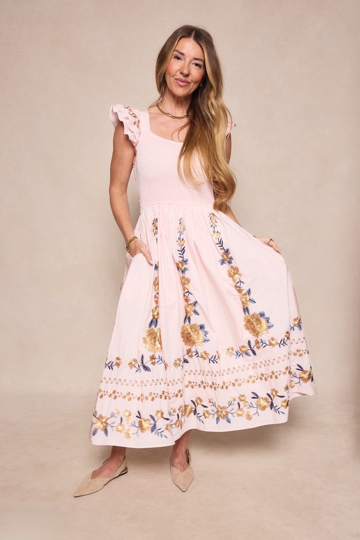 A woman models the Ivy City Co Hattie Dress in Embroidered Spring Floral, featuring ruffled cap sleeves and a maternity-friendly fit, styled with beige flats against a neutral background.