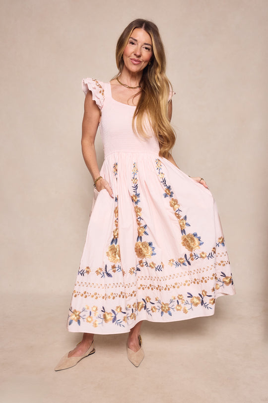 A woman models the Ivy City Co Hattie Dress in Embroidered Spring Floral, featuring ruffled cap sleeves and a maternity-friendly fit, styled with beige flats against a neutral background.