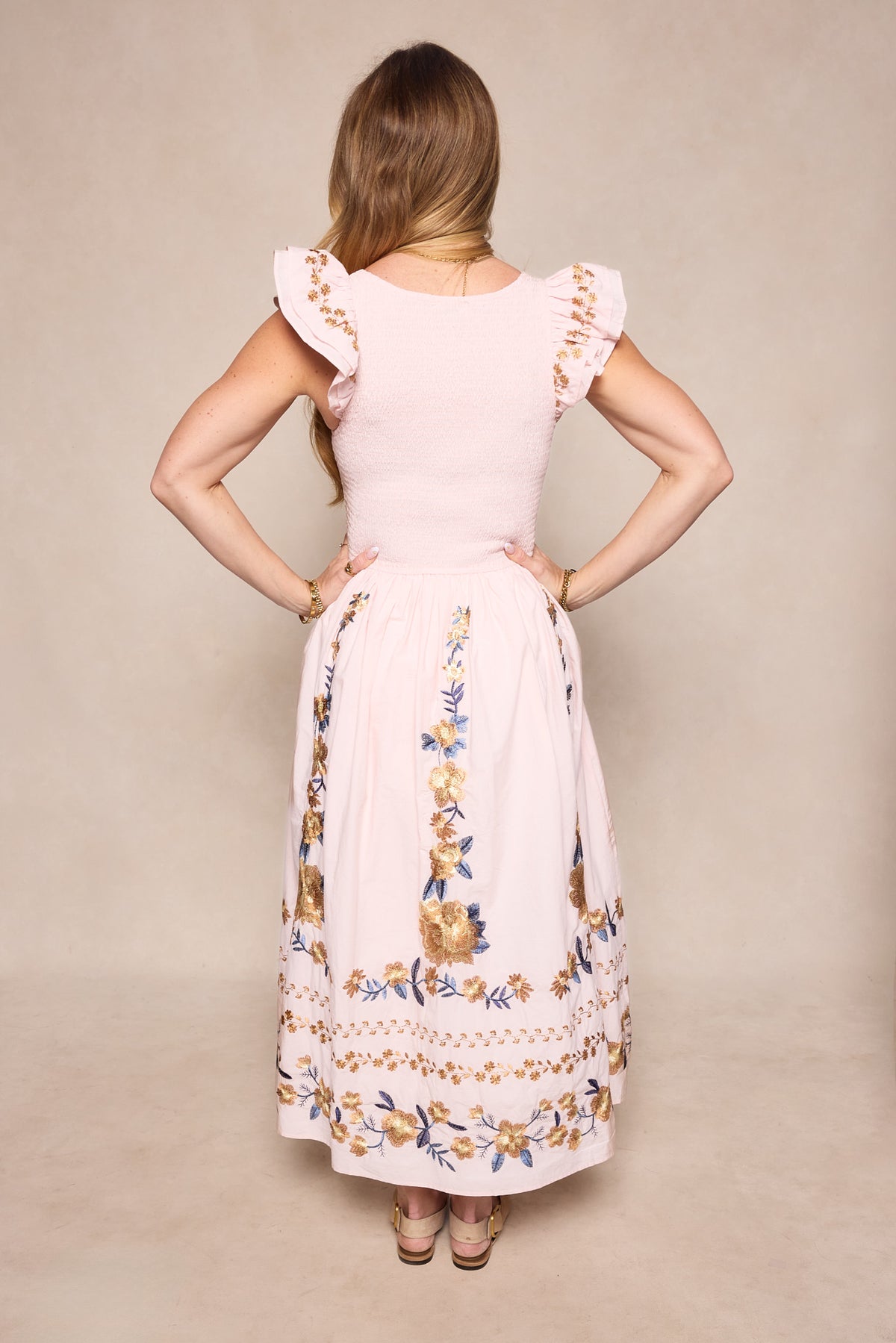 A woman with long, light brown hair stands facing away, hands on hips. She wears Ivy City Co’s Hattie Dress in Embroidered Spring Floral, featuring ruffled sleeves and a mid-calf skirt. The background is plain light beige.