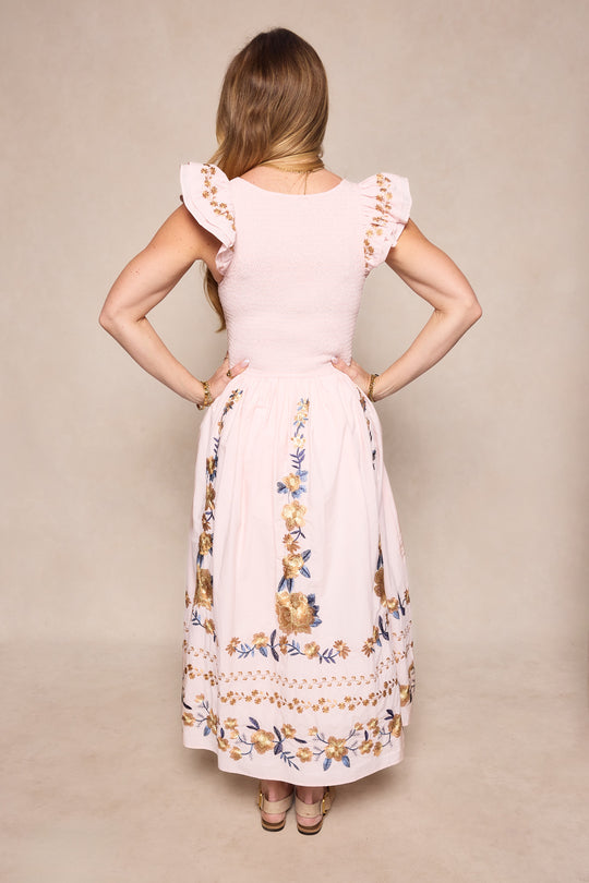 A woman with long, light brown hair stands facing away, hands on hips. She wears Ivy City Co’s Hattie Dress in Embroidered Spring Floral, featuring ruffled sleeves and a mid-calf skirt. The background is plain light beige.
