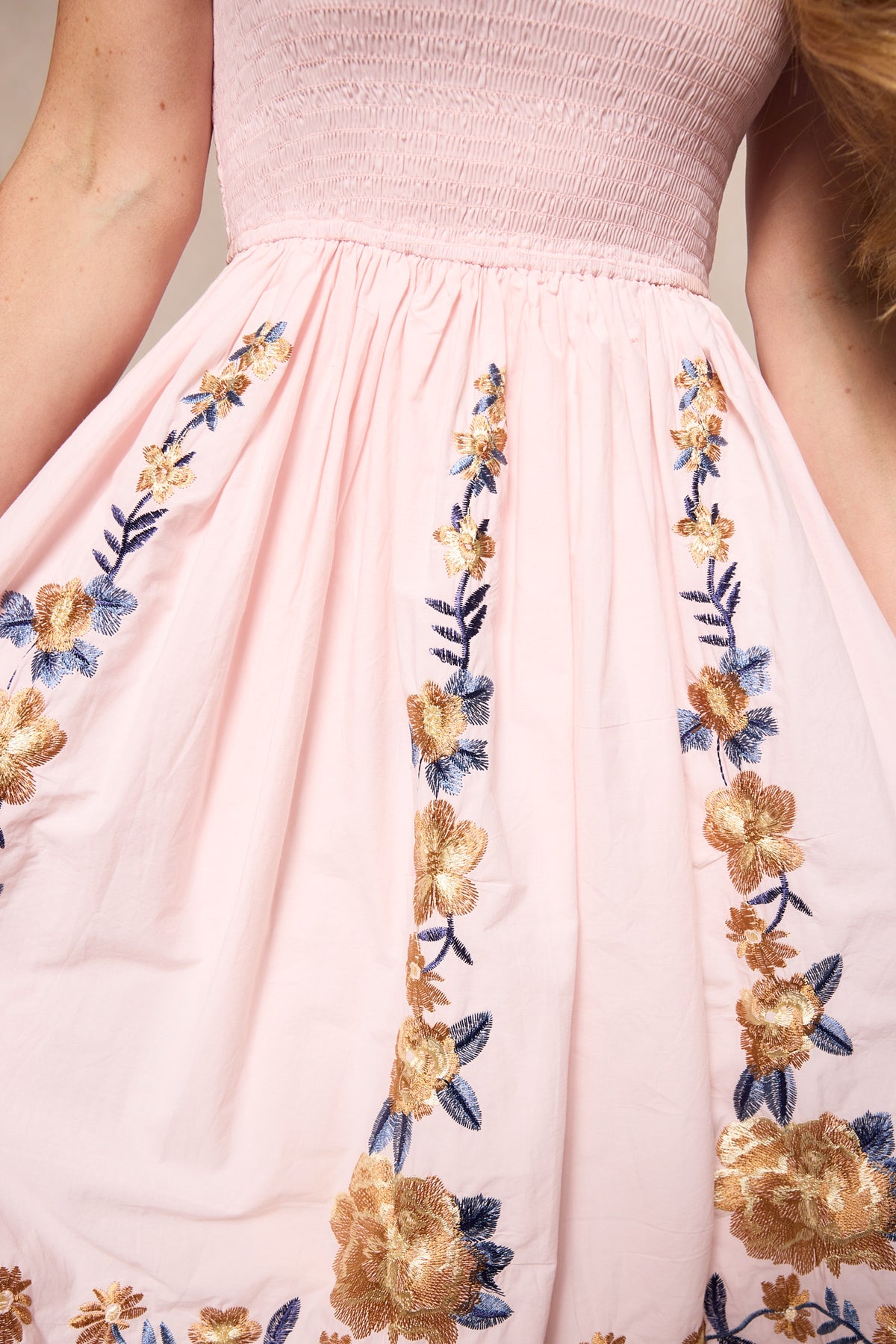 A woman wearing the Ivy City Co Hattie Dress in Embroidered Spring Floral—a light pink, maternity-friendly midi with a smocked bodice and gold and blue floral embroidery—gently holds out the sides of her dress.